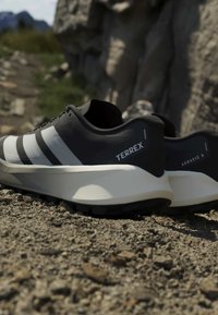Ασπρόμαυρα παπούτσια trail τρεξίματος Adidas Terrex Agravic 4, τοποθετημένα σε βραχώδες έδαφος με θολό εξωτερικό φόντο από πέτρες και πράσινο.