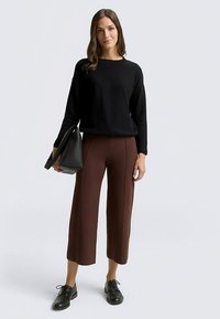 Maglione nero lavorato a maglia, pantaloni corti marroni con cuciture ricamate, scarpe nere allacciate e una borsa tote nera, su uno sfondo chiaro.