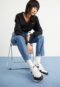 Hanorac negru Nike, blugi din denim albastru deschis, șosete albe și adidași Nike alb-negru, așezat pe un scaun argintiu împotriva unui fundal simplu.