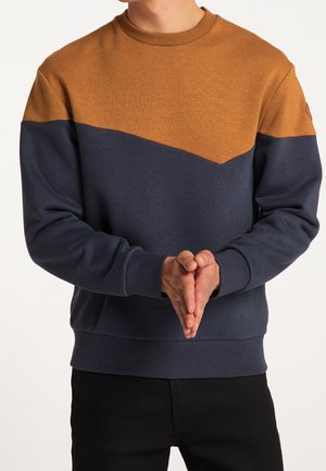 Mand iført en brun og marinefarvet sweatshirt med hænderne foldet foran torsoen, sorte bukser synlige nedenunder.