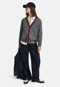 Grå texturerad cardigan med orange och marinblå V-ringning, svarta knappdetaljer, som bärs tillsammans med en vit skjorta, marinblå byxor och en basebollkeps.