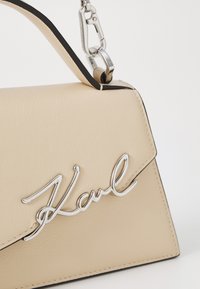 Beige leather handbag with silver hardware, featuring a prominent logo in cursive. Includes a top handle and contrasting black accents.