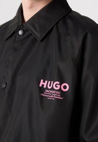 Czarna wodoodporna kurtka z wykładanym kołnierzem, z różowym logo i tekstem na klatce piersiowej. Gładka tekstura i zapięcie na napy.