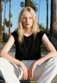 Blonde vrouw met los haar zit met gekruiste benen buiten, draagt een zwart mouwloos topje en witte broek, op de achtergrond palmbomen.