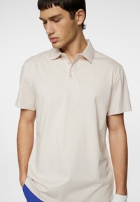 Ljust beige pikétröja i mjuk bomull, med klassisk krage, korta ärmar och en liten vit logotyp på bröstet.