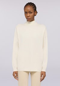 Maglione a collo alto crema con maniche lunghe, vestibilità ampia e texture liscia. Indossato con pantaloni beige abbinati, su uno sfondo chiaro.