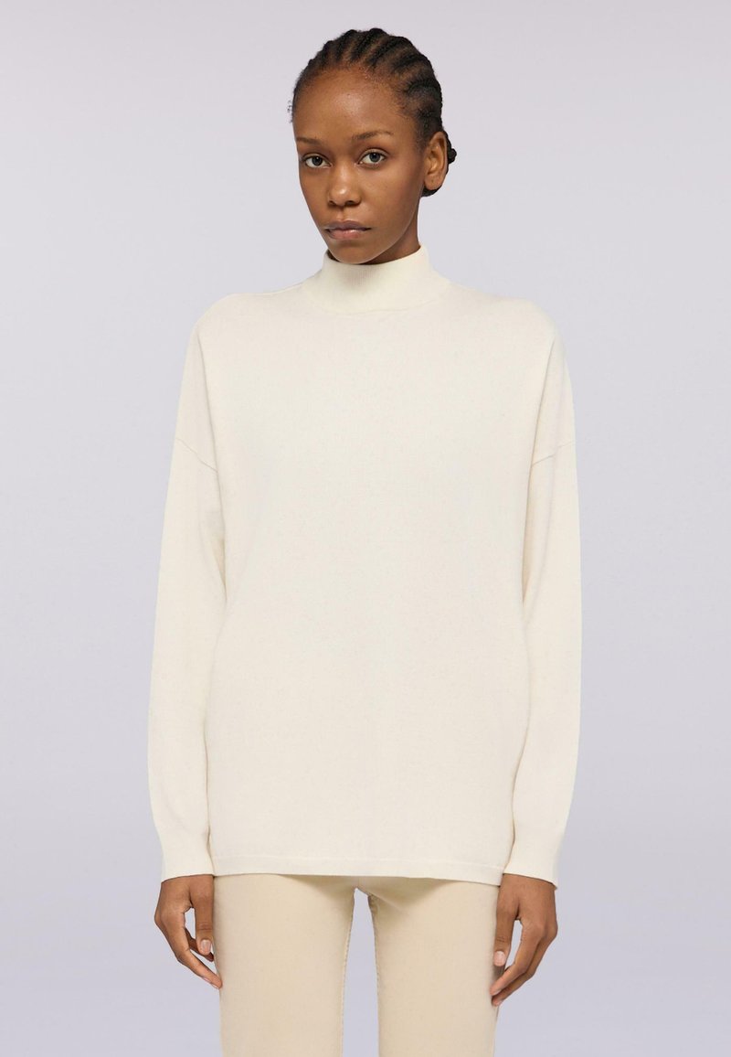 Maglione a collo alto crema con maniche lunghe, vestibilità ampia e texture liscia. Indossato con pantaloni beige abbinati, su uno sfondo chiaro.