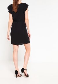 Robe noire avec des manches courtes en volant, une taille ceinturée et un ourlet à hauteur du genou. Portée avec des chaussures à talons hauts noires, dotées de brides de cheville.