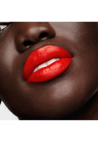 Un lápiz labial mate de un rojo brillante en labios carnosos, que contrasta con la piel oscura, presentando una textura suave y una forma bien definida con dientes blancos visibles.