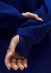 Felpa blu con polsini a coste; primo piano di una mano che afferra il tessuto mostrando la sua consistenza liscia e il design aderente, evidenziando la caratteristica del foro per il pollice.
