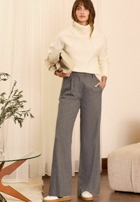 Krämfärgad polotröja, grå vida byxor med framveck, vita sneakers, texturerad tyg och minimalistisk clutch, placerat i ett ljust rum.