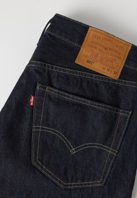 Σκούρη μπλε πίσω τσέπη τζιν Levi's με κίτρινη ραφή και καφέ δερμάτινη ετικέτα που δείχνει μέγεθος και λεπτομέρειες μάρκας.
