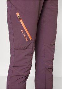 Pantalones outdoor Vaudé de color púrpura con un bolsillo con cremallera naranja, tejido texturizado y costuras reforzadas. Presenta un detalle del logo en la pierna.