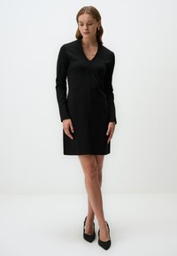 Robe noire à manches longues, décolleté en V et coupe droite. Texture lisse et design à hauteur des genoux. Portée avec des talons noirs.