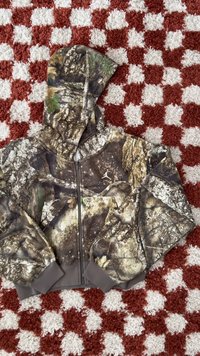 Hooded camouflage jacket with zipper and subtle leaf and bark patterns lying on a red and white checkered textured surface.