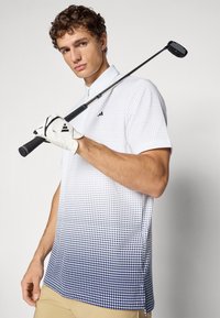 Mladý muž v bílém a námořnickém gradientovém golfovém tričku a rukavici, držící golfovou hůl přes rameno na jednotném pozadí.