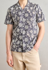 Homme portant une chemise à manches courtes avec un grand motif floral blanc sur fond sombre et un pantalon beige, une main dans la poche.