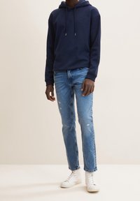 Námořnická mikina s klokaní kapsou, kombinovaná se světle modrými roztrhanými džíny a bílými vysokými teniskami, vytváří uvolněný vzhled.