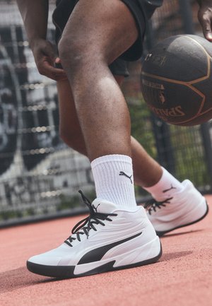 Zapatillas deportivas blancas con acentos negros, parte superior de malla texturizada, cordones negros y suela negra sobre una cancha de baloncesto roja.