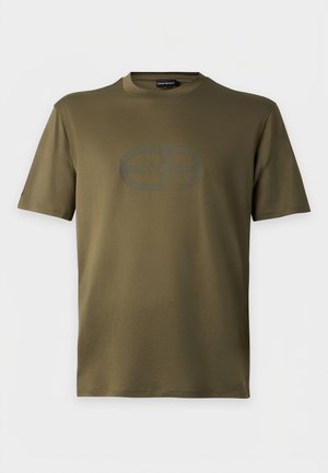 Homme portant des lunettes de soleil foncées, un t-shirt vert olive avec un logo, un pantalon gris foncé et des baskets beiges, debout devant un fond clair uni.