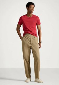 Vyras stovi su rankomis kišenėse, vilkėdamas raudoną marškinėlį, kreminės spalvos klostuotus kelnes, baltus sportinius batelius ir laikrodį prieš paprastą foną.