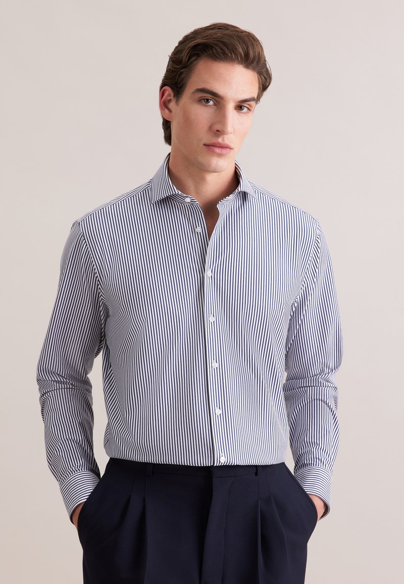 Jeune homme aux cheveux foncés portant une chemise à rayures verticales bleues et blanches boutonnée, rentrée dans un pantalon sombre plissé, les mains dans les poches.