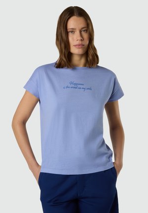 Donna con capelli castani fino alle spalle che indossa una maglietta azzurra con la scritta "La felicità è il vento nelle mie vele" e pantaloni blu scuro.