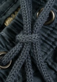 Cordón trenzado gris oscuro atado en un nudo, pasando a través de ojales metálicos, con un fondo de tela negra texturizada que presenta líneas verticales.