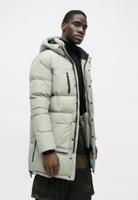 Ανοιχτό γκρι φουσκωτό μπουφάν με κουκούλα, που διαθέτει τσέπες με φερμουάρ, κλείσιμο με φερμουάρ μπροστά και υφή με κουβάρες.