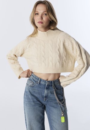 Giovane donna in un maglione corto lavorato a maglia a treccia color crema con zip sulle maniche, jeans blu a vita alta e una catena con un accessorio a tag verde.