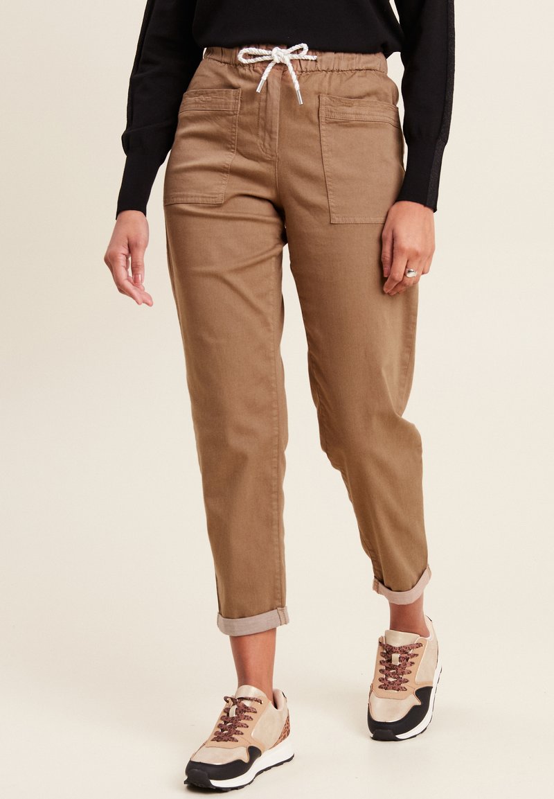 Beige, taps toelopende broek met een elastische tailleband en witte trekkoord. Voorzien van twee zijzakken en opgerolde manchetten. Draagt bij beige sneakers.