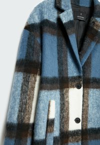 Abrigo de mezcla de lana a cuadros en azul, blanco y marrón. Presenta un cuello con muescas, botones negros y bolsillos laterales con una textura suave.