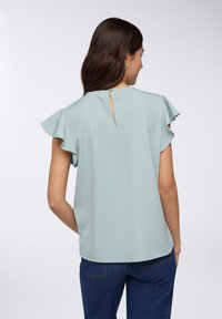 Blusa verde chiaro con maniche corte a volant e dettaglio a occhiello sulla schiena, realizzata in un tessuto morbido, abbinata a jeans blu scuro.