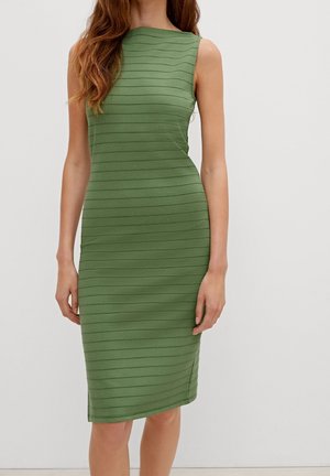 Femme portant une robe verte sans manches, ajustée, longueur genou, avec des rayures horizontales discrètes, debout devant un fond clair uni.