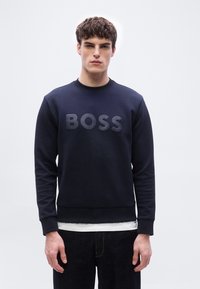 Νέος άνδρας φοράει μπλε σκούρο φούτερ με την επιγραφή "BOSS" στην μπροστινή πλευρά, λευκό εσωτερικό μπλουζάκι εμφανές και σκουρόχρωμο παντελόνι, πάνω σε απλό φόντο.