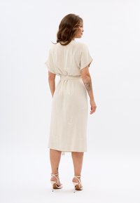 Beiges Leinenkleid mit tailliertem Schnitt und kurzen Ärmeln, mit einem knielangen Saum und dezenten bestickten Details. Trägt leichte Absätze.