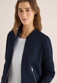 Femme aux cheveux bruns mi-longs portant une veste zippée bleu marine sur une chemise blanche, sur un fond uni.