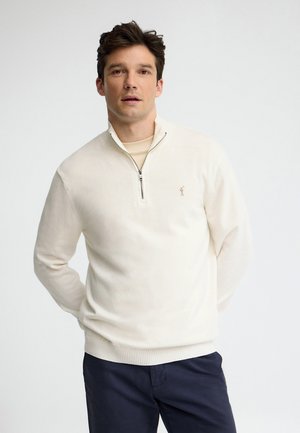 Mann trägt einen cremefarbenen Pullover mit Viertel-Reißverschluss und marineblaue Hose, steht mit verschränkten Händen hinter dem Rücken vor einem schlichten, hellen Hintergrund.