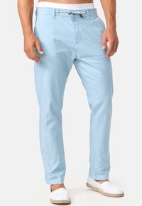 Pantaloni azzurri chiari, slim-fit, realizzati in un tessuto traspirante. Presentano una vita con cordoncino e due tasche laterali, abbinati a scarpe bianche senza lacci.