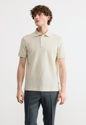 Νεαρός άνδρας με σγουρά μαλλιά, φορώντας υφασμάτινο μπλουζάκι πόλο σε εκρού απόχρωση και σκούρο γκρι παντελόνι, στέκεται μπροστά σε απλό ανοιχτόχρωμο φόντο.