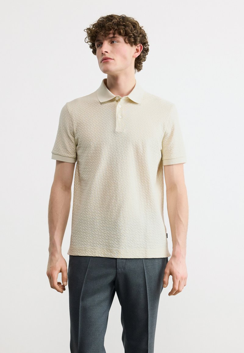 Νεαρός άνδρας με σγουρά μαλλιά, φορώντας υφασμάτινο μπλουζάκι πόλο σε εκρού απόχρωση και σκούρο γκρι παντελόνι, στέκεται μπροστά σε απλό ανοιχτόχρωμο φόντο.