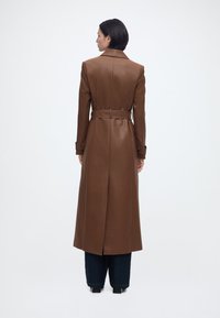 Γυναίκα με κοντά σκούρα μαλλιά που φορά ένα μακρύ καφέ δερμάτινο παλτό με ζώνη, σκούρο παντελόνι και μαύρα παπούτσια, στέκεται με την πλάτη στραμμένη σε λευκό φόντο.