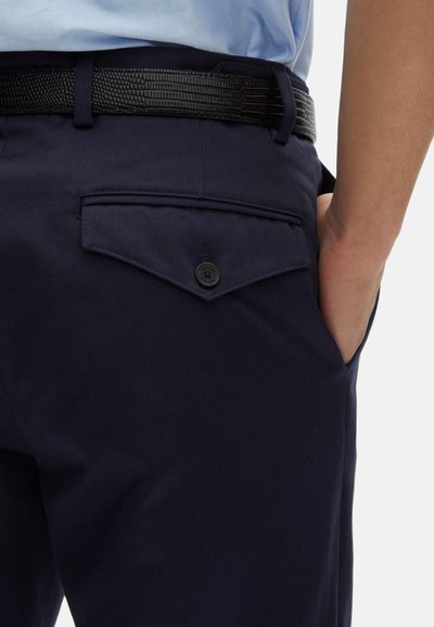 Marineblauwe broek met een gladde textuur, voorzien van een achterzak met een klep en knoopsluiting. Zwart textuurceintuur zichtbaar rond de taille.