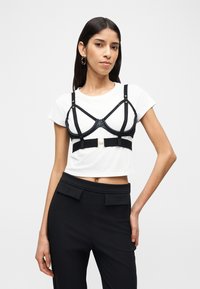 Γυναίκα που φοράει λευκό κοντό t-shirt με μαύρο λουρί στήθους πάνω από αυτό και μαύρο ψηλόμεσο παντελόνι με λεπτομέρειες με πατιλέτα.