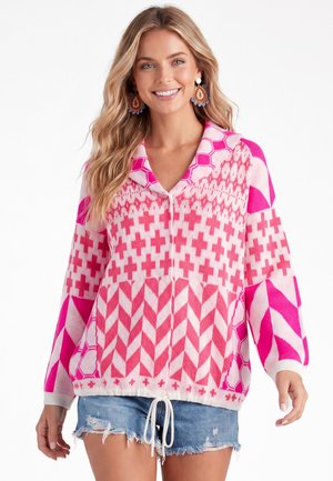 Smilende kvinde iført farverig pink mønstret strikket sweater med krave og blå denimshorts, accessoriseret med store perleøreringe.