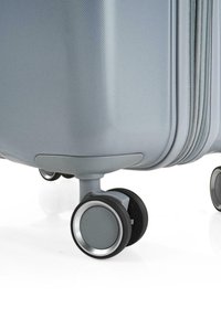 Maleta rígida gris con una superficie texturizada, tres ruedas suaves con acentos plateados y un diseño aerodinámico.