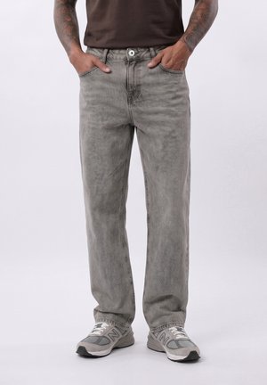 Person trägt verblasste graue Jeans mit geradem Bein, Hände in den Taschen, graue Sneakers und ein dunkelbraunes Hemd, gezeigt von der Taille bis zu den Füßen.