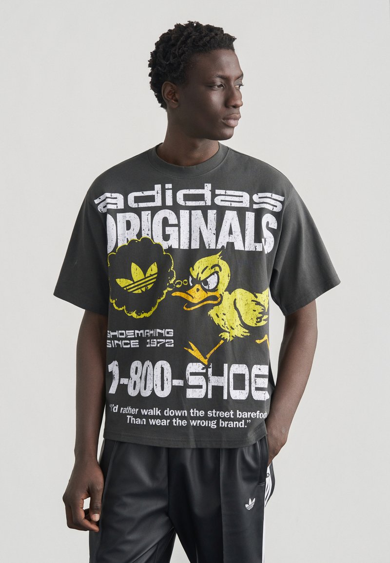 Άνδρας που φοράει μαύρο oversized T-shirt Adidas Originals με κίτρινο γραφικό θυμωμένης πάπιας και κείμενο, συνδυασμένο με μαύρο παντελόνι φόρμας Adidas.