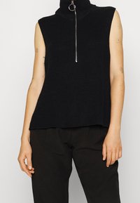 Černý bezrukávový rolák vyrobený z žebrované pleteniny, s kovovým zipem a uvolněným střihem na lemu.