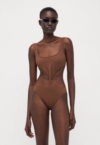 Brown textured one-piece swimsuit with a square neckline and cut-out design. Model wears dark sunglasses.